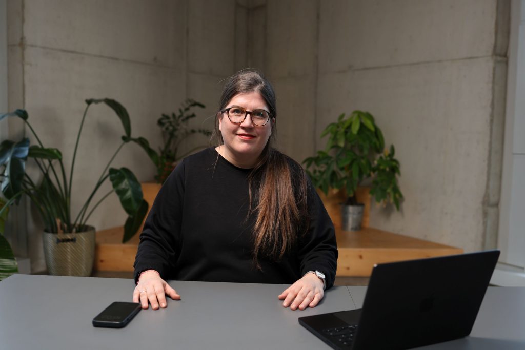 Anne-Marie Bernhard, eine Person mit langen Haaren und Brille sitzt an einem Tisch mit Notebook aufgeklappt und Smartphone.
Im Hintergrund Pflanzen.